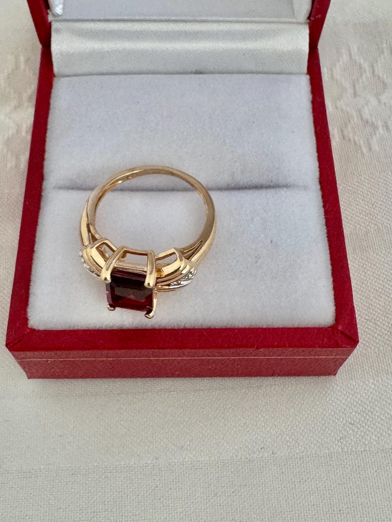 Op de afbeelding: Een gouden ring met een rechthoekige, dieprode edelsteen. De ring is gezet in een gouden band met kleine accentstenen. De ring wordt gepresenteerd in een rode sieradendoos met een witte binnenkant.