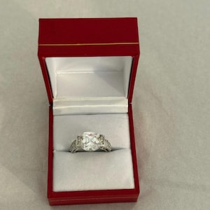 May include: A silver ring with a large, square, clear stone is displayed inside a red jewelry box with a white interior. The ring has smaller stones on the band. The box is open, revealing the ring.