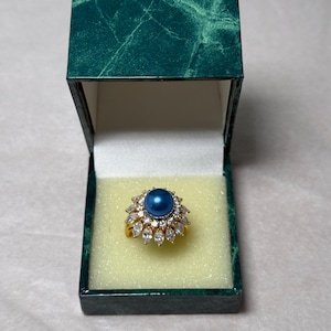 May include: A gold ring with a large, round, dark blue center stone surrounded by smaller clear stones. The ring is displayed in a green marble-patterned jewelry box with a yellow interior.