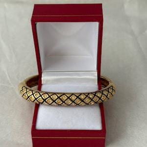 May include: A gold-toned bangle bracelet with a black and diamond-shaped pattern, set with small, clear gemstones. The bracelet rests inside an open, red jewelry box with a white interior, suggesting a gift or presentation.