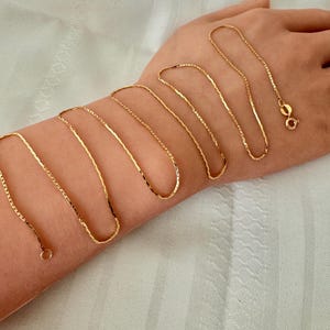 May include: A gold-toned chain necklace featuring a delicate box link design. The chain is composed of small, square-shaped links connected in a continuous, flexible strand. It includes a circular spring ring clasp at one end for secure fastening.