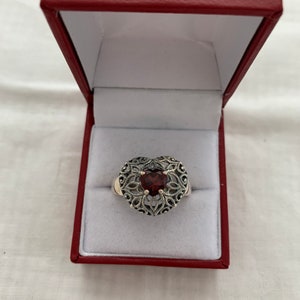 May include: A silver ring with a heart-shaped design and a red gemstone in the center. The ring is in a red box.