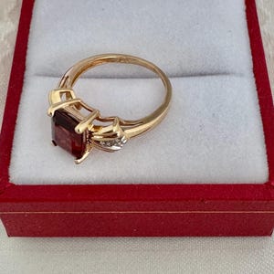 Op de afbeelding: Een gouden ring met een grote, rechthoekige, donkerrode edelsteen. De ring heeft een decoratieve zetting met kleine accentstenen. Gepresenteerd in een rode sieradendoos met een witte binnenkant.