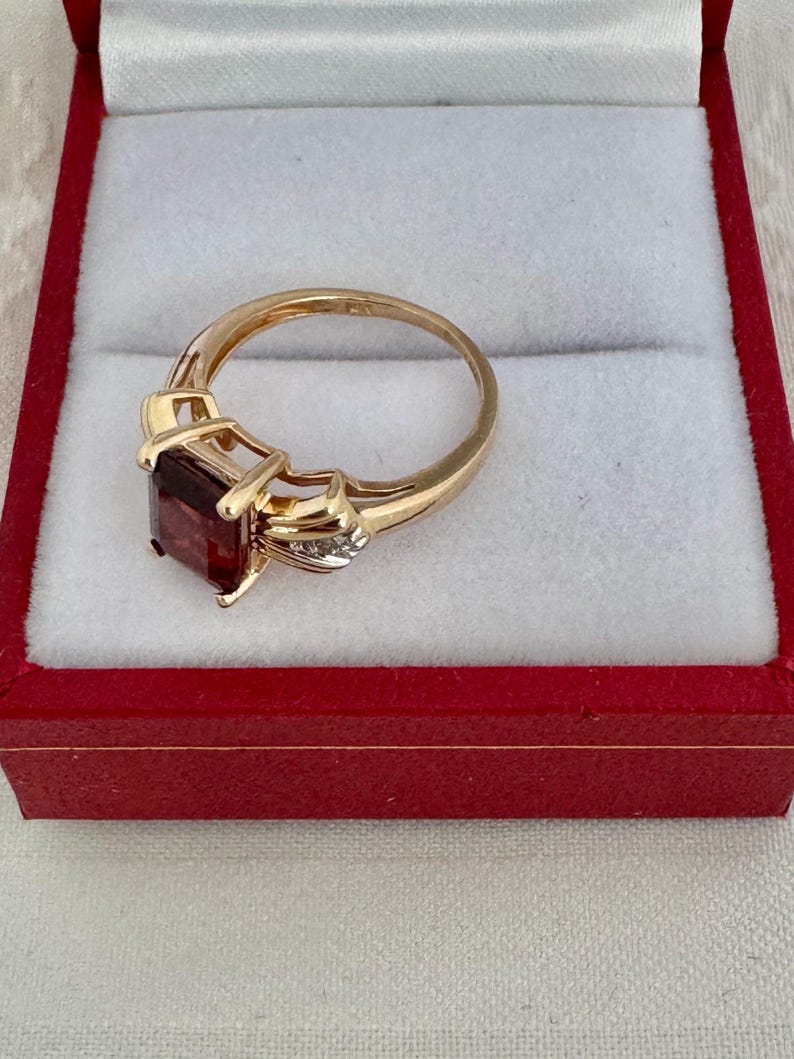 Op de afbeelding: Een gouden ring met een grote, rechthoekige, donkerrode edelsteen. De ring heeft een decoratieve zetting met kleine accentstenen. Gepresenteerd in een rode sieradendoos met een witte binnenkant.