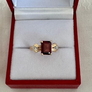 Op de afbeelding: Een gouden ring met een rechthoekige, dieprode edelsteen. De ring heeft kleine diamantaccenten aan weerszijden van de edelsteen. De ring wordt gepresenteerd in een rode sieradendoos met een wit interieur.