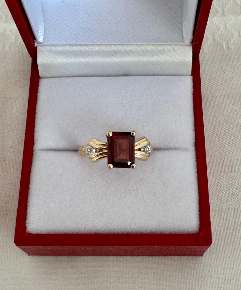 Op de afbeelding: Een gouden ring met een rechthoekige, dieprode edelsteen. De ring heeft kleine diamantaccenten aan weerszijden van de edelsteen. De ring wordt gepresenteerd in een rode sieradendoos met een wit interieur.