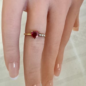 May include: A gold ring featuring a central heart-shaped red gemstone and three small clear accent stones on one side of the band. The ring is worn on a finger, showcasing the polished gold metal band and the vibrant red color of the heart stone.