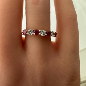 May include: A gold band ring featuring alternating round-cut gemstones. The ring showcases a pattern of red rubies and clear, sparkling stones. The ring is displayed on a finger, highlighting the jewelry's design and color contrast.