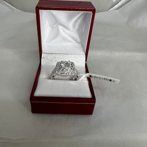 May include: A silver ring with a square-shaped center stone, set in a decorative filigree design. The ring is displayed in a red jewelry box with a white satin interior. A price tag is attached to the box.