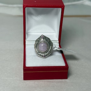 May include: A silver-toned ring with a light pink oval stone, set in a decorative frame with small red accents. The ring is displayed in a red jewelry box with a white interior, suggesting a gift or presentation setting.