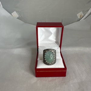 May include: A silver ring with a large oval green gemstone surrounded by smaller stones. The ring is in a red velvet box.