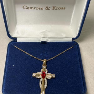 May include: A gold-toned cross pendant necklace with a red center stone and clear accent stones, displayed in a blue velvet jewelry box. The box lid is open, revealing the brand name "Camrose & Kross".