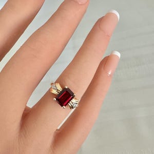 Op de afbeelding: Een gouden ring met een rechthoekige, dieprode edelsteen als middelpunt. De ring heeft een decoratief ontwerp met kleinere, heldere edelstenen aan weerszijden van de hoofdsteen. De ring zit om een vinger.