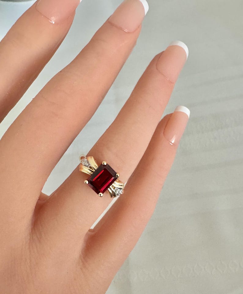 Op de afbeelding: Een gouden ring met een rechthoekige, dieprode edelsteen als middelpunt. De ring heeft een decoratief ontwerp met kleinere, heldere edelstenen aan weerszijden van de hoofdsteen. De ring zit om een vinger.