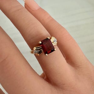 Op de afbeelding: Een gouden ring met een rechthoekige, dieprode edelsteen. De ring heeft kleine accentdiamanten aan weerszijden van de edelsteen. De ring zit om een vinger.