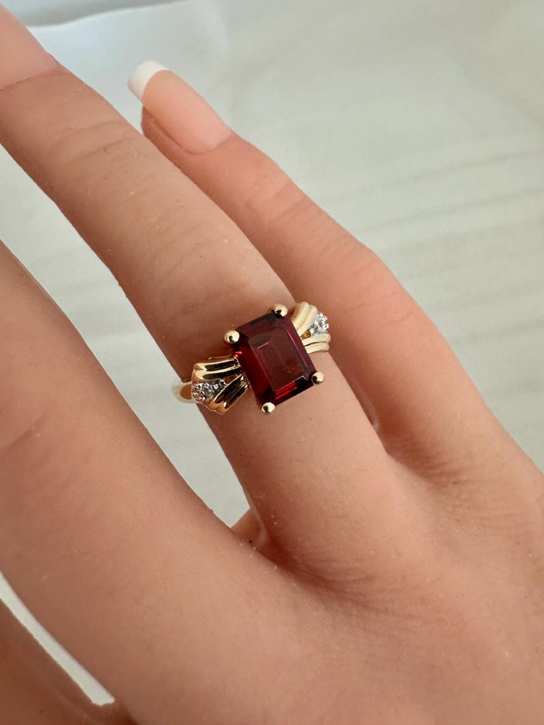 Op de afbeelding: Een gouden ring met een rechthoekige, dieprode edelsteen. De ring heeft kleine accentdiamanten aan weerszijden van de edelsteen. De ring zit om een vinger.