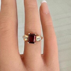 Op de afbeelding: Een gouden ring met een grote, rechthoekige, dieprode edelsteen. De ring heeft een gouden band met decoratieve accenten en kleine, heldere stenen. De ring wordt op een vinger getoond. De belangrijkste steen is ongeveer 1 cm lang.