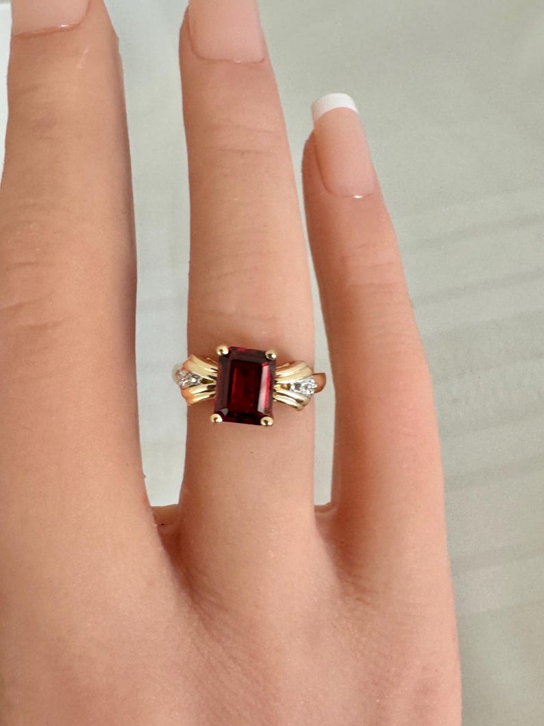 Op de afbeelding: Een gouden ring met een grote, rechthoekige, dieprode edelsteen. De ring heeft een gouden band met decoratieve accenten en kleine, heldere stenen. De ring wordt op een vinger getoond. De belangrijkste steen is ongeveer 1 cm lang.