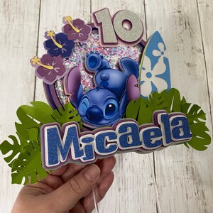May include: A blue Stitch character cake topper with a surfboard, flowers, and green leaves. The topper also includes the number 10 and the name Micaela.