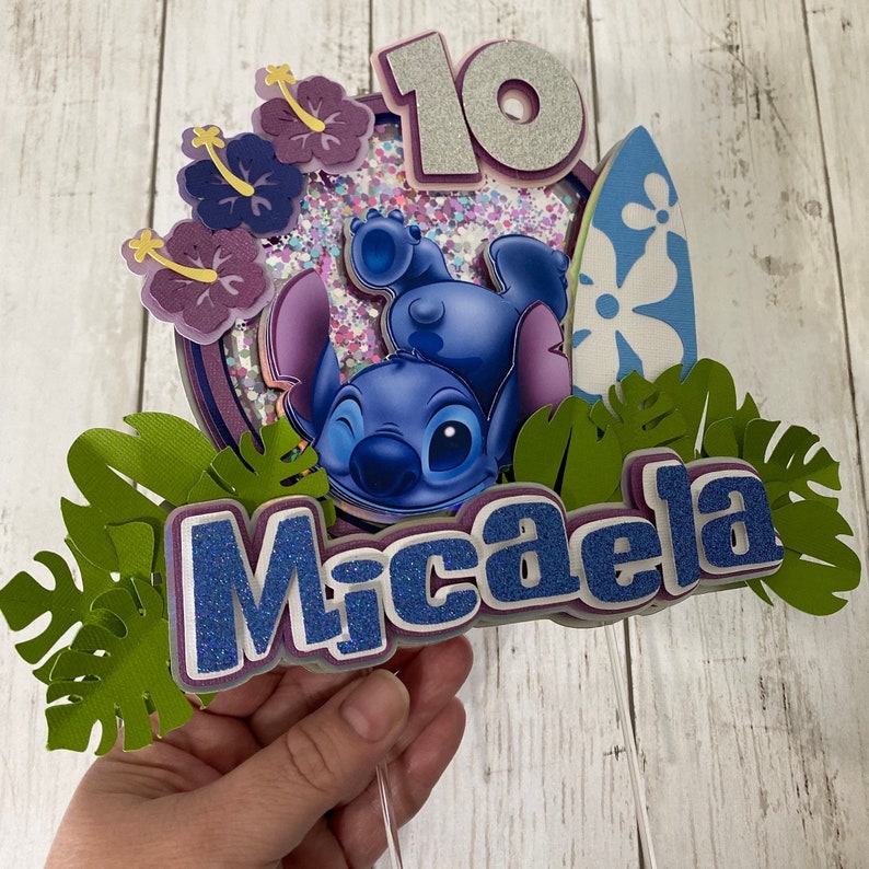 May include: A blue Stitch character cake topper with a surfboard, flowers, and green leaves. The topper also includes the number 10 and the name Micaela.