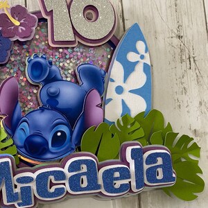 May include: A blue and purple cake topper featuring Stitch from Lilo & Stitch, a surfboard, and the name "Licaela" in blue glitter letters. The cake topper is decorated with glitter and a purple flower.