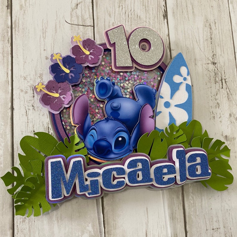 May include: A blue Stitch character from Lilo & Stitch is featured on a cake topper with a surfboard, purple flowers, and green leaves. The number 10 and the name Micaela are also included on the topper.