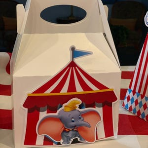 Puede incluir: Caja de regalo de cartón blanco con una carpa de circo a rayas rojas y blancas y una imagen de dibujos animados de Dumbo el elefante. La caja tiene un asa en la parte superior.