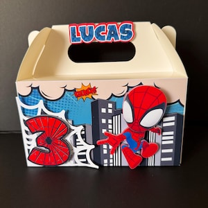 May include: A cream-colored party favor box featuring a Spider-Man theme. The box has a handle and is decorated with a cityscape, a red number 3, and the name "LUCAS" in blue letters. A Spider-Man figure is also on the box.