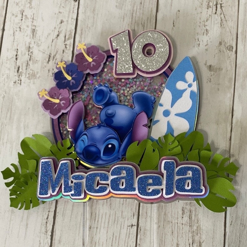 May include: A blue Stitch character cake topper with a surfboard, flowers, and the name Micaela in glitter letters. The number 10 is also included in glitter.