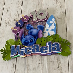 May include: A blue Stitch character from Lilo & Stitch is on a cake topper with a surfboard, green leaves, purple flowers, and the name Micaela. The number 10 is on the cake topper with glitter.
