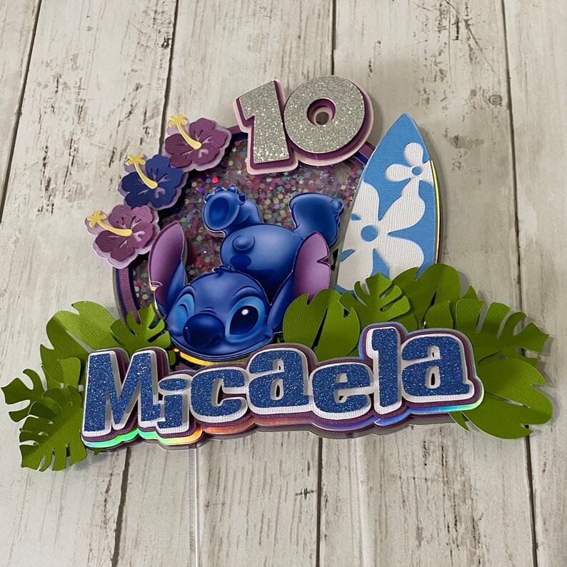 May include: A blue Stitch character from Lilo & Stitch is on a cake topper with a surfboard, green leaves, purple flowers, and the name Micaela. The number 10 is on the cake topper with glitter.