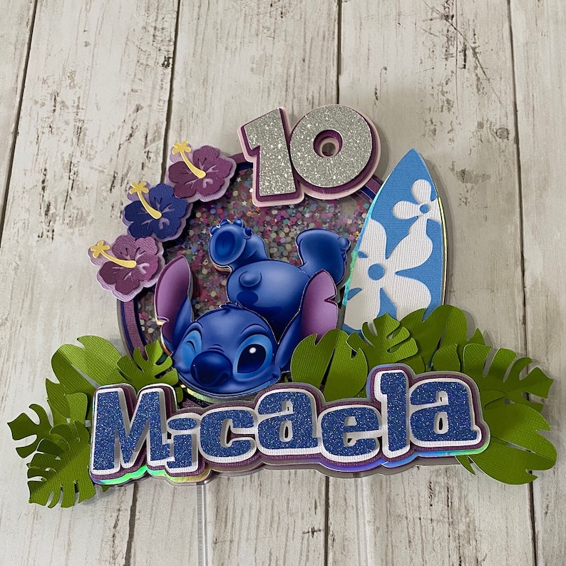 May include: A blue Stitch character cake topper with a surfboard, purple flowers, green leaves, and the name Micaela in blue glitter letters. The number 10 is also included in silver glitter.