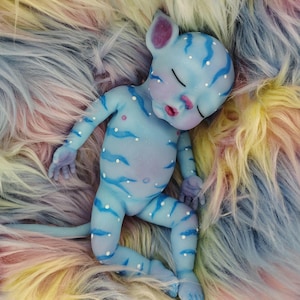 May include: A blue fantasy doll with a cat-like face and pointed ears, lying on a multicolored faux fur surface. The doll has blue stripes and white dots on its body, with closed eyes and a small tail. The fur is a mix of pastel colors.