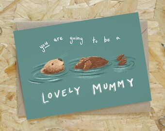 Puede incluir: Tarjeta de felicitación con ilustración de una nutria en el agua y el texto "you are going to be a LOVELY MUMMY" sobre fondo turquesa. La tarjeta está sobre un sobre marrón, con un lápiz y clips.