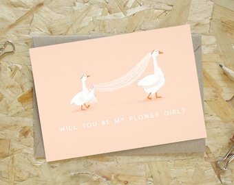 Puede incluir: Una tarjeta de felicitación rectangular con fondo color melocotón claro que muestra una ilustración de dos gansos blancos, uno con un velo sostenido por el otro. El texto "WILL YOU BE MY FLOWER GIRL?" está impreso en blanco en la parte inferior.