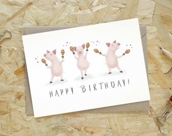 Puede incluir: Tarjeta de cumpleaños con tres cerdos rosas tocando maracas. El texto "HAPPY BIRTHDAY!" está escrito debajo. La tarjeta está sobre una superficie de madera, con un lápiz y una pinza.