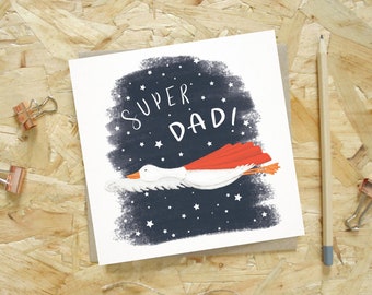 Puede incluir: Tarjeta de felicitación con la frase "SUPER DAD!" en blanco sobre un fondo azul oscuro con estrellas blancas. Un ganso blanco con capa roja vuela por la tarjeta. Un lápiz y clips están sobre una superficie de madera.