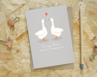 Peut inclure: Carte de vœux de mariage avec un fond gris, représentant deux oies blanches et un cœur rouge. Le texte dit "To my love on our wedding day". Un crayon et des pinces à reliure sont sur la surface en bois.