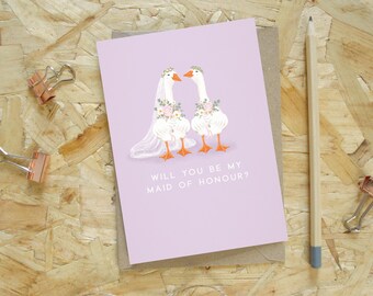 Puede incluir: Una tarjeta de felicitación de color lila con el texto «WILL YOU BE MY MAID OF HONOUR?» presenta dos gansos de dibujos animados con coronas de flores y velos. La tarjeta está sobre un sobre marrón, con un lápiz y dos clips de encuadernación color cobre cerca.