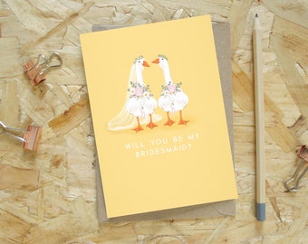 Puede incluir: Una tarjeta amarilla con el texto "WILL YOU BE MY BRIDESMAID?" muestra dos gansos vestidos de boda, con coronas florales y ramos. También se ven un sobre marrón, un lápiz y dos clips de encuadernación de cobre.