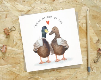 Puede incluir: Una tarjeta de felicitación cuadrada con una ilustración de dos patos sosteniendo tazas blancas, con el texto YOU'RE MY CUP OF TEA y un pequeño corazón rojo encima. Los patos son marrones y verdes con patas naranjas, de pie juntos.