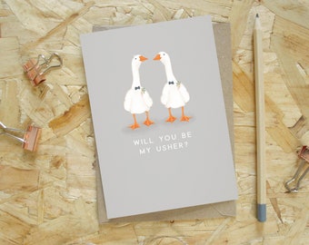 Puede incluir: Tarjeta de felicitación gris con dos gansos de dibujos animados con pajaritas y detalles florales. El texto "WILL YOU BE MY USHER?" está impreso debajo. La tarjeta está sobre un sobre marrón, con un lápiz y dos clips cerca.
