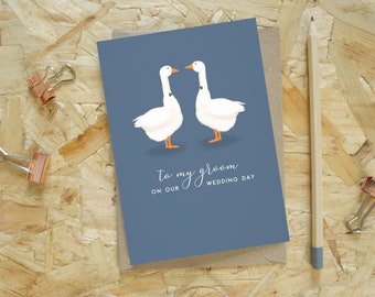 Peut inclure: Carte de vœux avec un fond bleu représentant deux oies blanches avec des nœuds papillon noirs. Le texte "To my groom ON OUR WEDDING DAY" est écrit en script. La carte est sur une enveloppe marron, avec un crayon et des trombones.