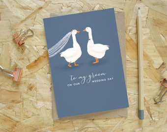 Puede incluir: Una tarjeta de boda con fondo azul presenta dos gansos blancos, uno con velo y otro con pajarita. El texto dice "To my groom on our wedding day". Un lápiz y clips de papel están sobre la superficie de madera.