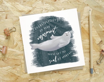 Puede incluir: Tarjeta de felicitación de compromiso con una ilustración de foca blanca. El texto "Felicidades por tu compromiso" y "Tienes el sello de aprobación" sobre un fondo azul oscuro.