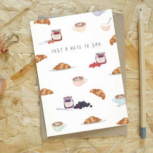 May include: A card with a watercolour design of croissants, jam jars, and coffee cups. The text "JUST A NOTE TO SAY" is printed on the front. A pencil and binder clips are on the wooden surface.