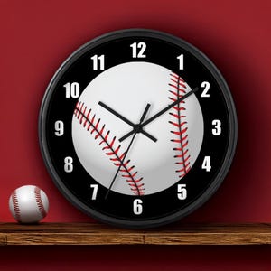 May include: A black-framed wall clock with a baseball design. The clock face features a white baseball with red stitching and black numbers. A baseball sits on a wooden shelf below the clock. The clock hands are black.