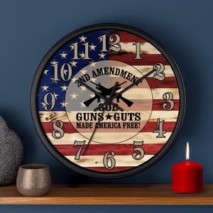 May include: A round wall clock with a black frame, featuring an American flag design. The clock face includes the text "2ND AMENDMENT, GOD, GUNS, GUTS, MADE AMERICA FREE!" and numerals. A red candle is lit on a wooden shelf.