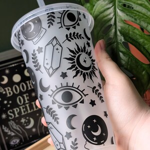 K&ouml;nnte beinhalten: Ein durchsichtiger Kunststoffbecher mit Deckel und Strohhalm, verziert mit schwarzen Himmels- und mystischen Symbolen. Das Design umfasst Monde, Sterne, Augen, Kristalle und Sonnen. Der Becher wird in der N&auml;he eines Buches mit dem Titel "Book of Spells" gehalten.