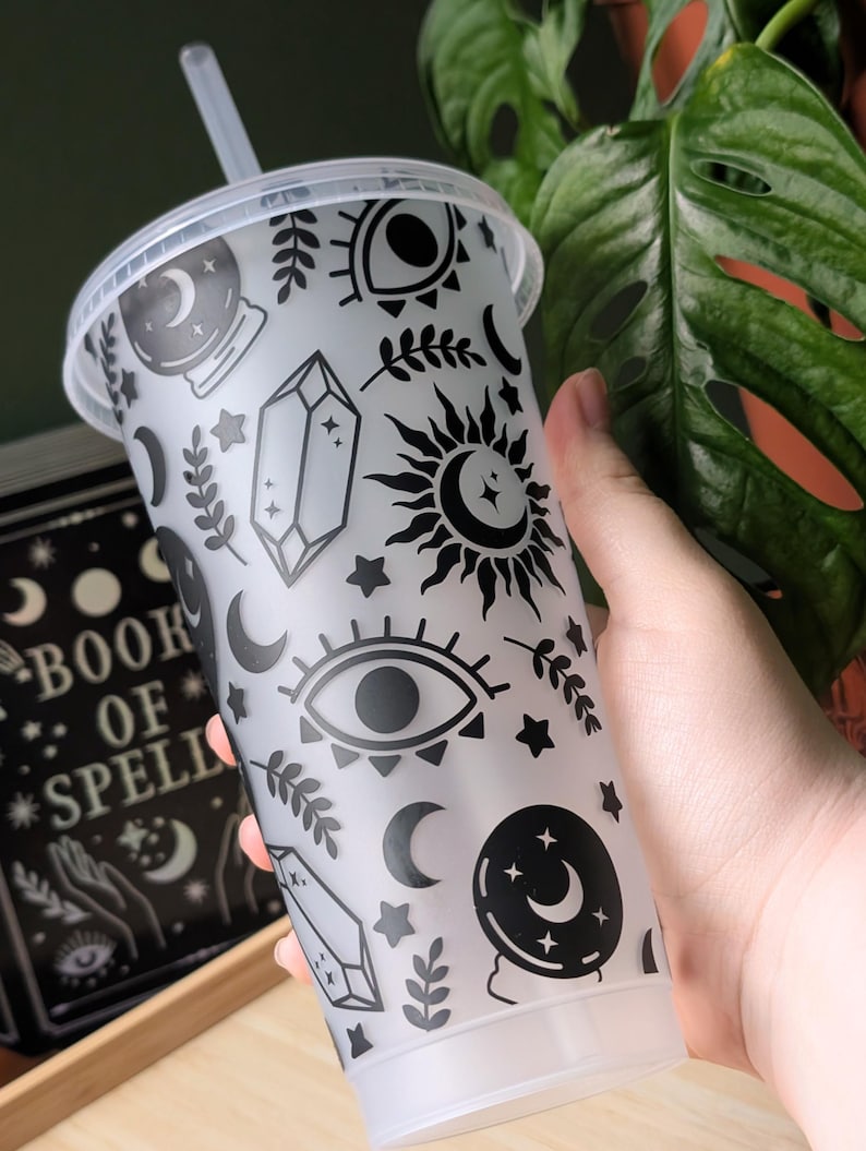 K&ouml;nnte beinhalten: Ein durchsichtiger Kunststoffbecher mit Deckel und Strohhalm, verziert mit schwarzen Himmels- und mystischen Symbolen. Das Design umfasst Monde, Sterne, Augen, Kristalle und Sonnen. Der Becher wird in der N&auml;he eines Buches mit dem Titel "Book of Spells" gehalten.